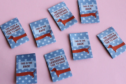Blue sausage dog sewing labels by The Cosy Club Handmade, double-sided fabric tags featuring playful dog design.