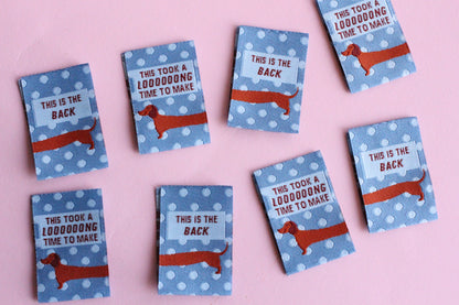 Blue sausage dog sewing labels by The Cosy Club Handmade, double-sided fabric labels with playful dog design for crafts and clothing.