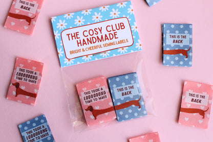 Pink and blue sausage dog sewing labels by The Cosy Club Handmade, double-sided fabric labels featuring cute dog designs.