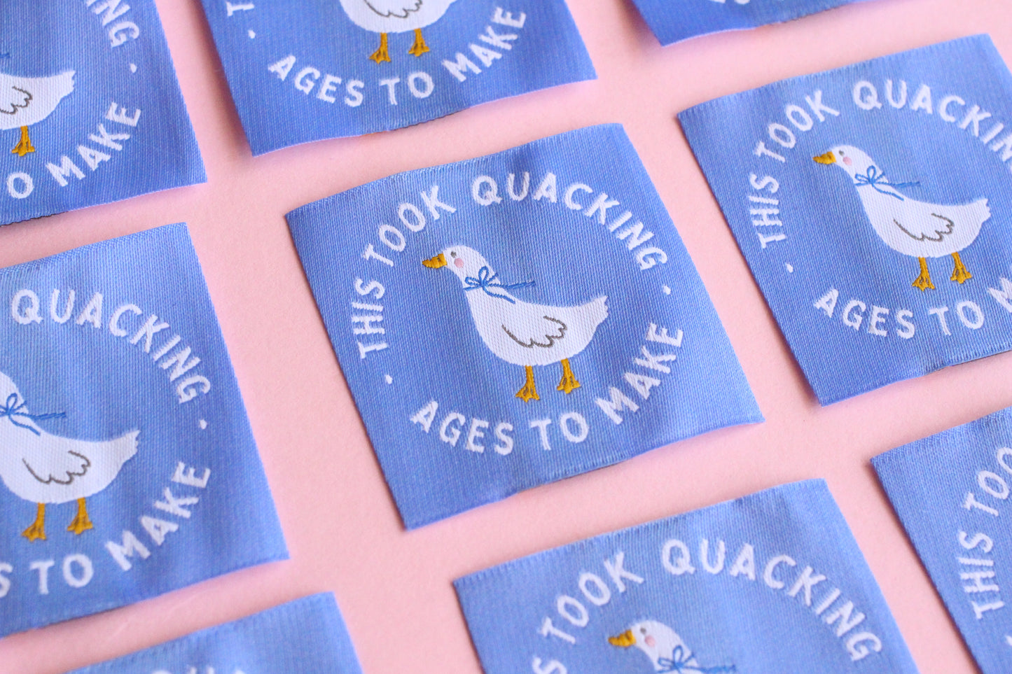 White goose sewing labels with "This Took Quacking Ages To Make" text from The Cosy Club Handmade, laid out on a neutral background.