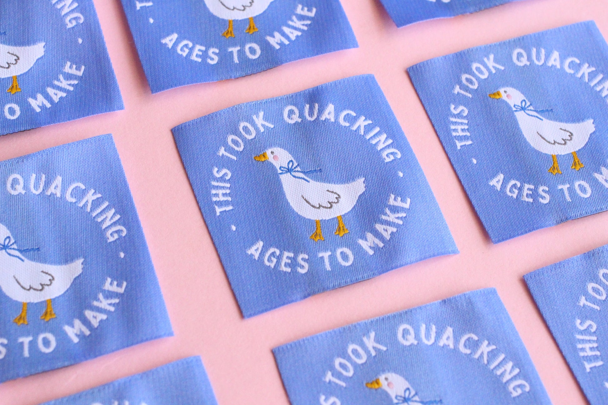 White goose sewing labels with "This Took Quacking Ages To Make" text from The Cosy Club Handmade, laid out on a neutral background.