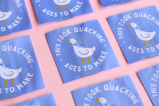 White goose sewing labels with "This Took Quacking Ages To Make" text from The Cosy Club Handmade, laid out on a neutral background.